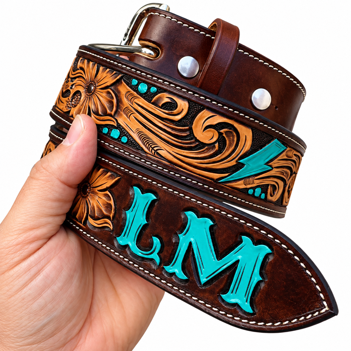 A hand holding a custom tooled leather belt. The belt is dark brown with intricate orange-brown floral and scroll tooling, turquoise accents, and the letters 'LM' tooled into the leather.