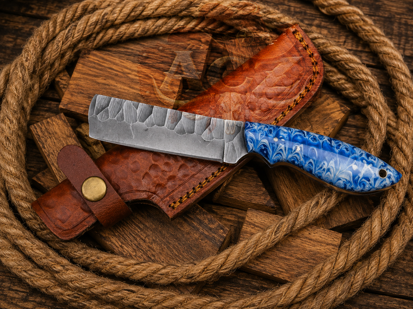 A fixed-blade knife with a hammered silver blade and a blue marbled handle, displayed diagonally on a brown leather sheath over a rustic wooden background with a coiled rope.