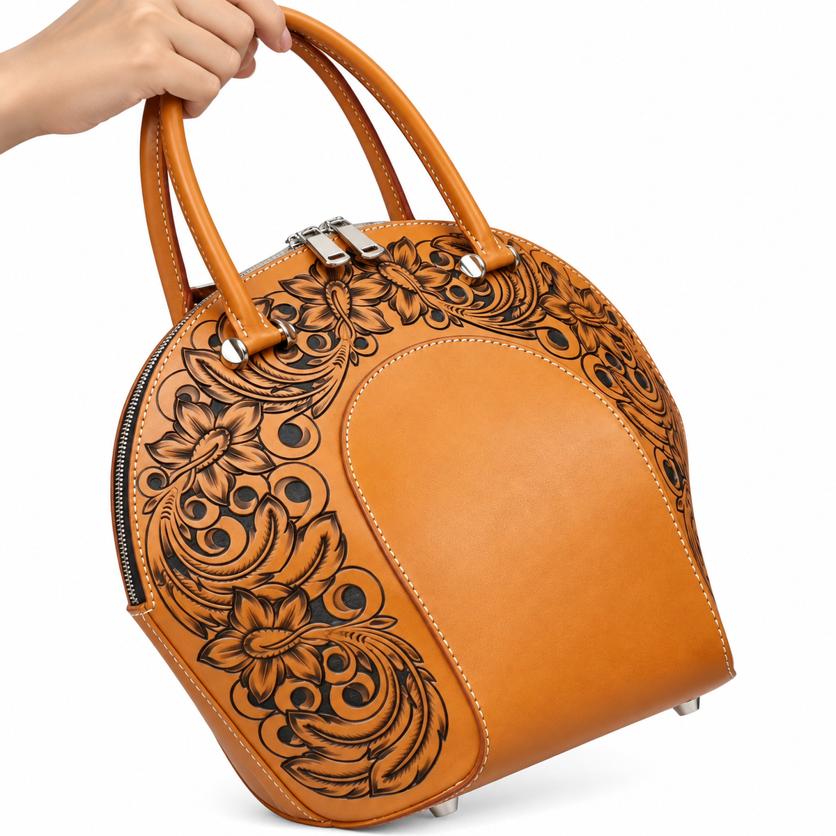 A hand holds the top handle of a tan leather dome-shaped handbag with black floral tooling on the sides, shown against a plain white background.
