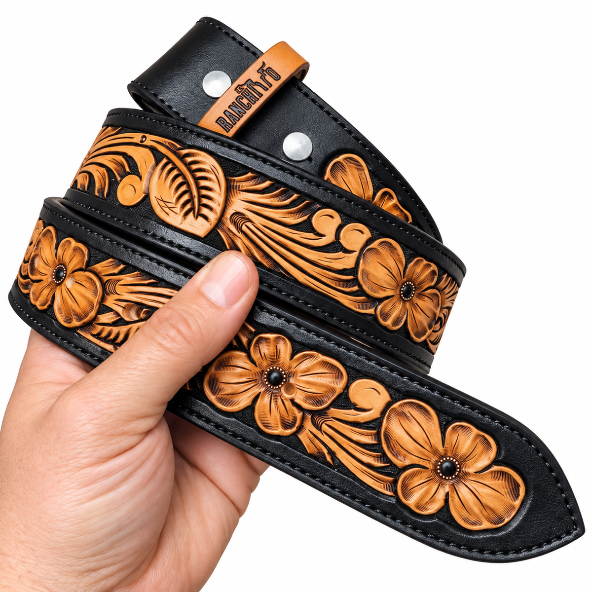 A hand holds a black leather belt with intricate brown tooled floral and leaf patterns. The belt features silver metal rivets and a small tan leather brand tab stamped 'Buckeye'.