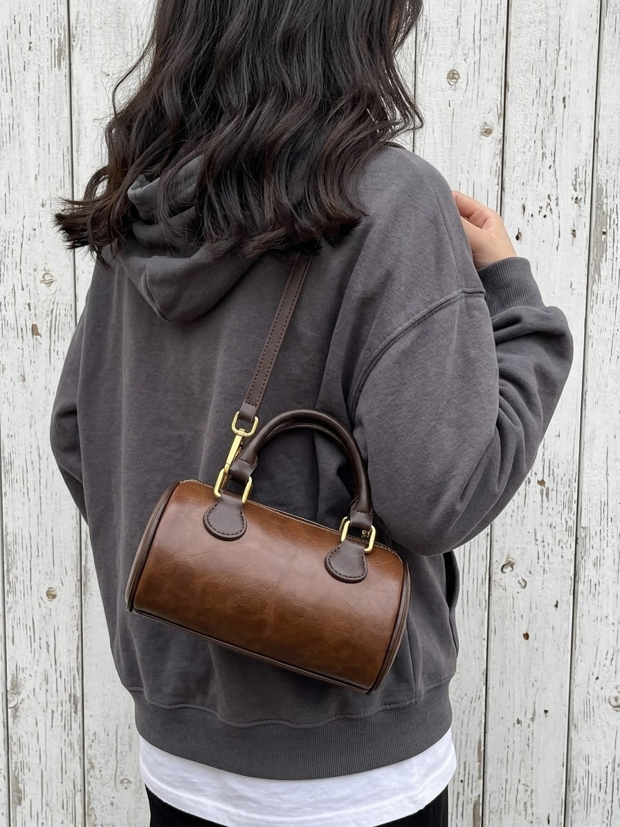 A person wearing a dark gray hoodie and white t-shirt is shown from behind, carrying a brown leather top-handle barrel bag with gold-tone hardware against a white wood plank wall.