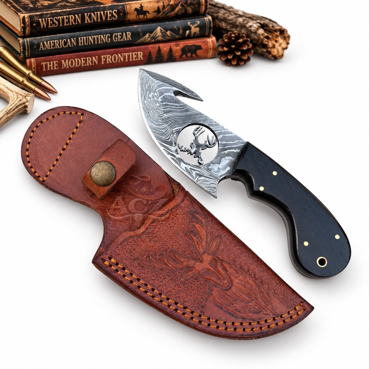 A fixed-blade knife with a patterned silver blade and black handle is displayed beside a brown leather sheath with an engraved deer design. The scene is styled on a white background with rustic props including books, wood, and bullets.