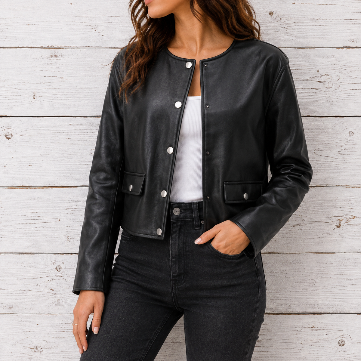 A woman wearing a black faux-leather cropped jacket with silver snap buttons, styled over a white top and black jeans