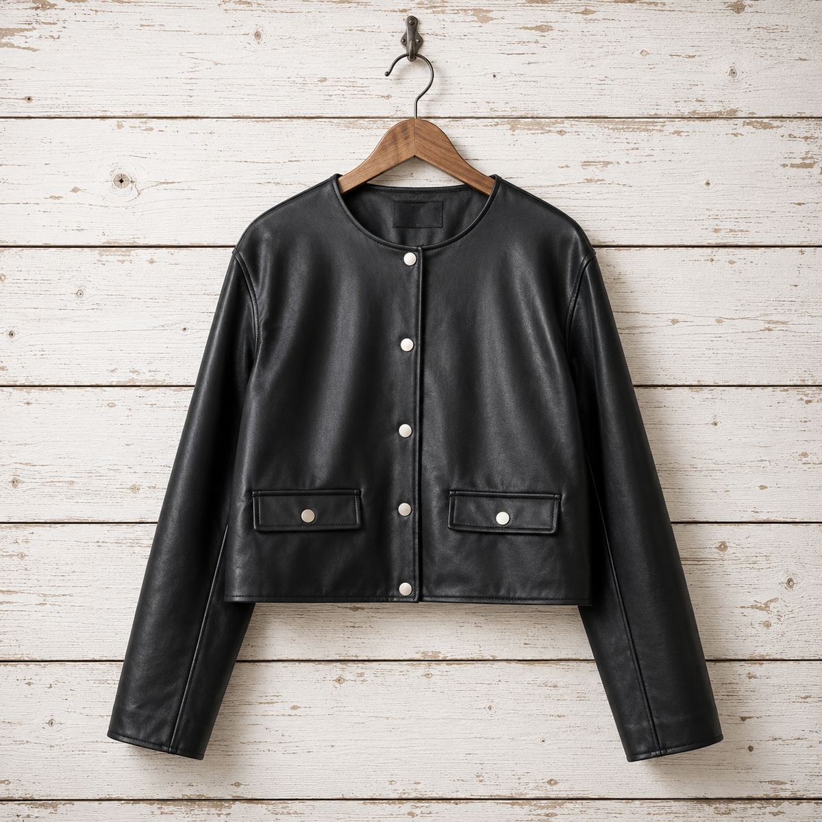 A black faux leather cropped jacket with a round neckline, snap-button front, and two front flap pockets, hanging on a wooden hanger 