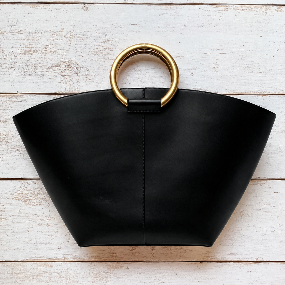 A black leather tote bag with a wide, structured fan shape and a large circular gold metal ring handle, displayed flat on a white wood plank background.