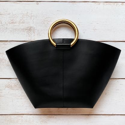 A black leather tote bag with a wide, structured fan shape and a large circular gold metal ring handle, displayed flat on a white wood plank background.