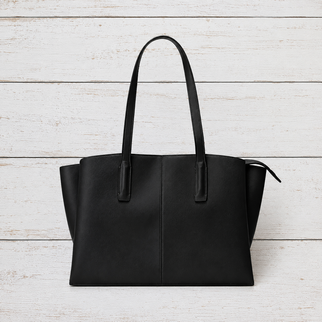 A black leather tote bag with long shoulder straps, displayed flat against a white-washed wooden plank background.