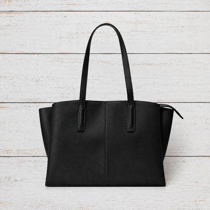 A black leather tote bag with long shoulder straps, displayed flat against a white-washed wooden plank background.