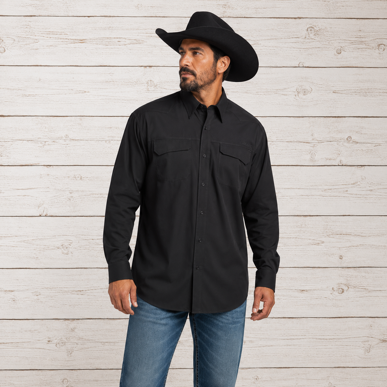 A man wearing a black long-sleeve western-style button-up shirt, blue jeans, and a black cowboy hat