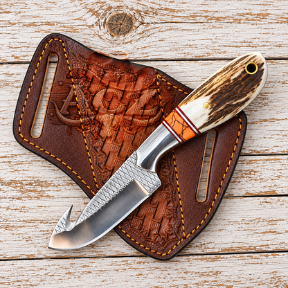 A custom hand-forged knife with a curved hook blade, textured steel, and a carved bone handle with an orange accent, displayed on a brown embossed leather sheath over a whitewashed wooden background.