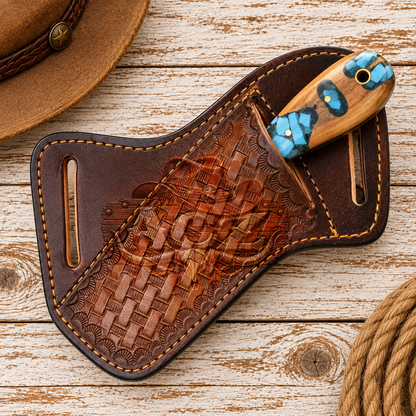 A brown leather knife sheath with a carved wooden handle featuring blue and gold inlay, displayed on a rustic whitewashed wooden surface with a hat and rope partially visible.
