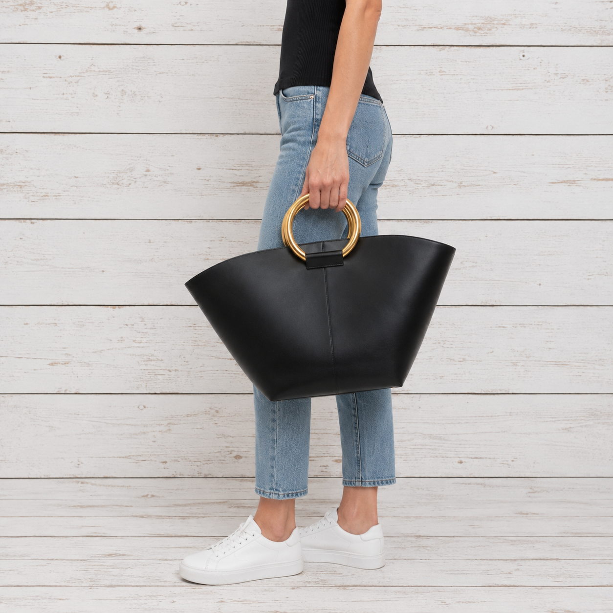 A person wearing light blue jeans and white sneakers holds a large black leather tote bag with a circular gold handle, standing against a white wood plank wall.