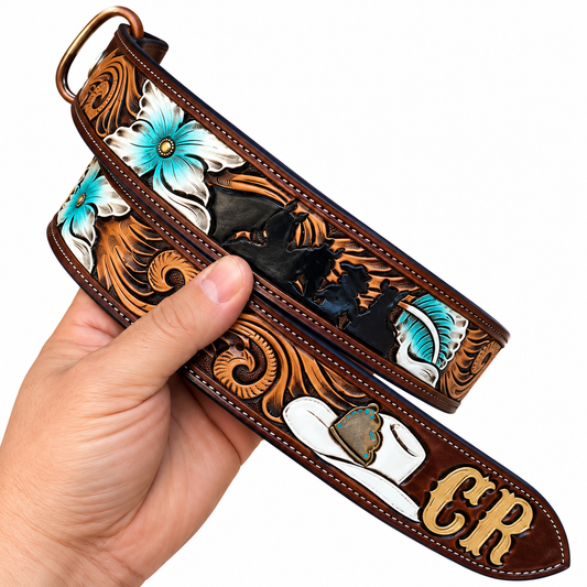 A hand holds a tooled leather belt featuring detailed western-style carving with turquoise blue flowers, black horses, white and brown boots, and gold monogram letters on a white background.