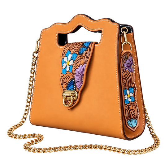 An orange leather shoulder bag with a top handle, gold chain strap, and colorful floral embroidery on the front flap, displayed on a rustic white-washed wooden background.
