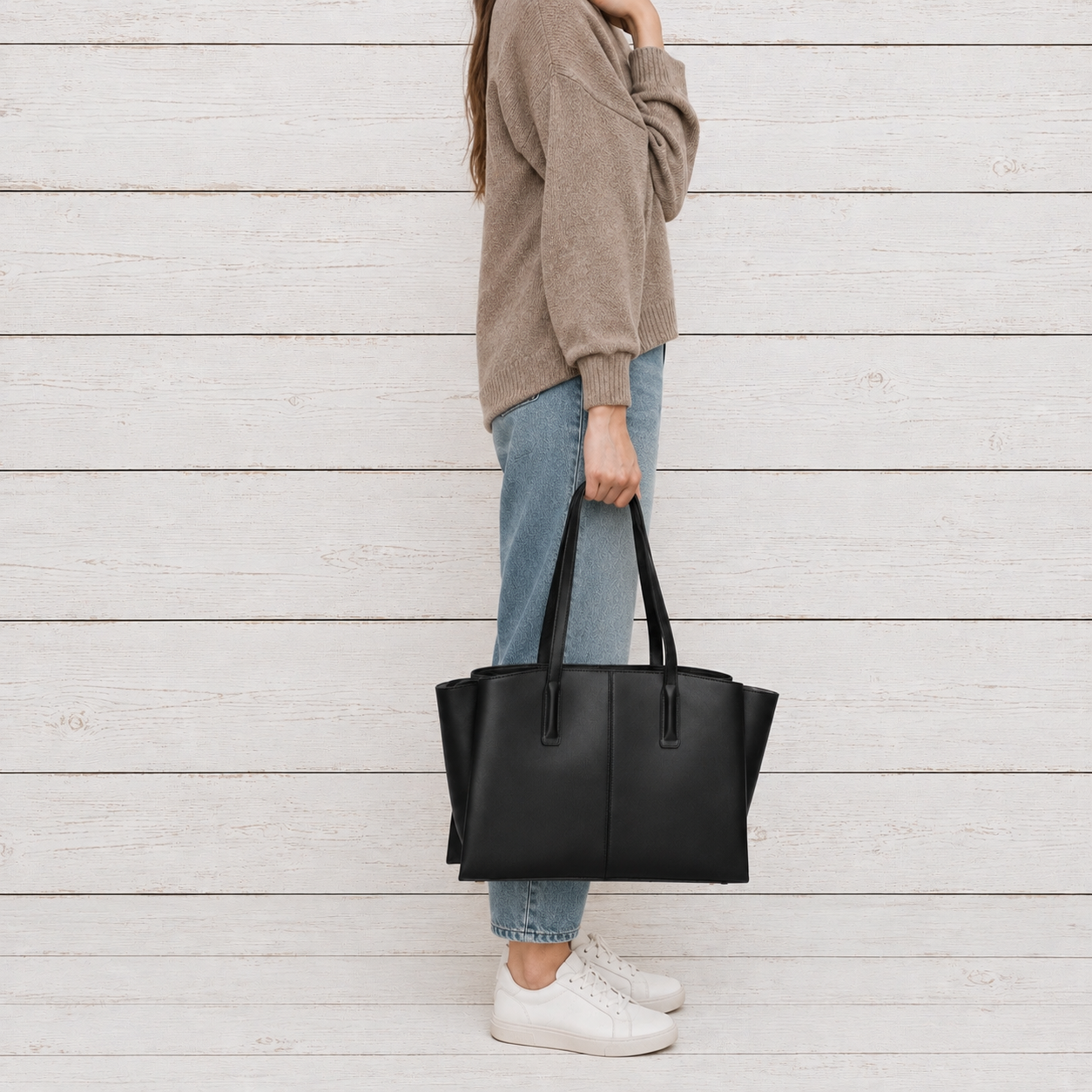 A person wearing a beige sweater, blue jeans, and white sneakers holds a large black leather tote bag against a white wood plank background.