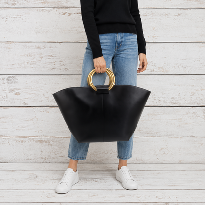 large black leather tote bag with two gold-tone circular handles against a white wood plank background.