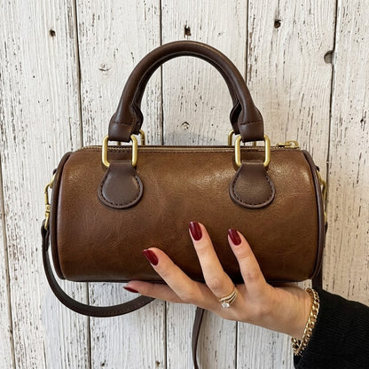 A brown leather top-handle barrel bag with gold-tone hardware, held by a hand with dark red nail polish against a rustic white-painted wood plank background.