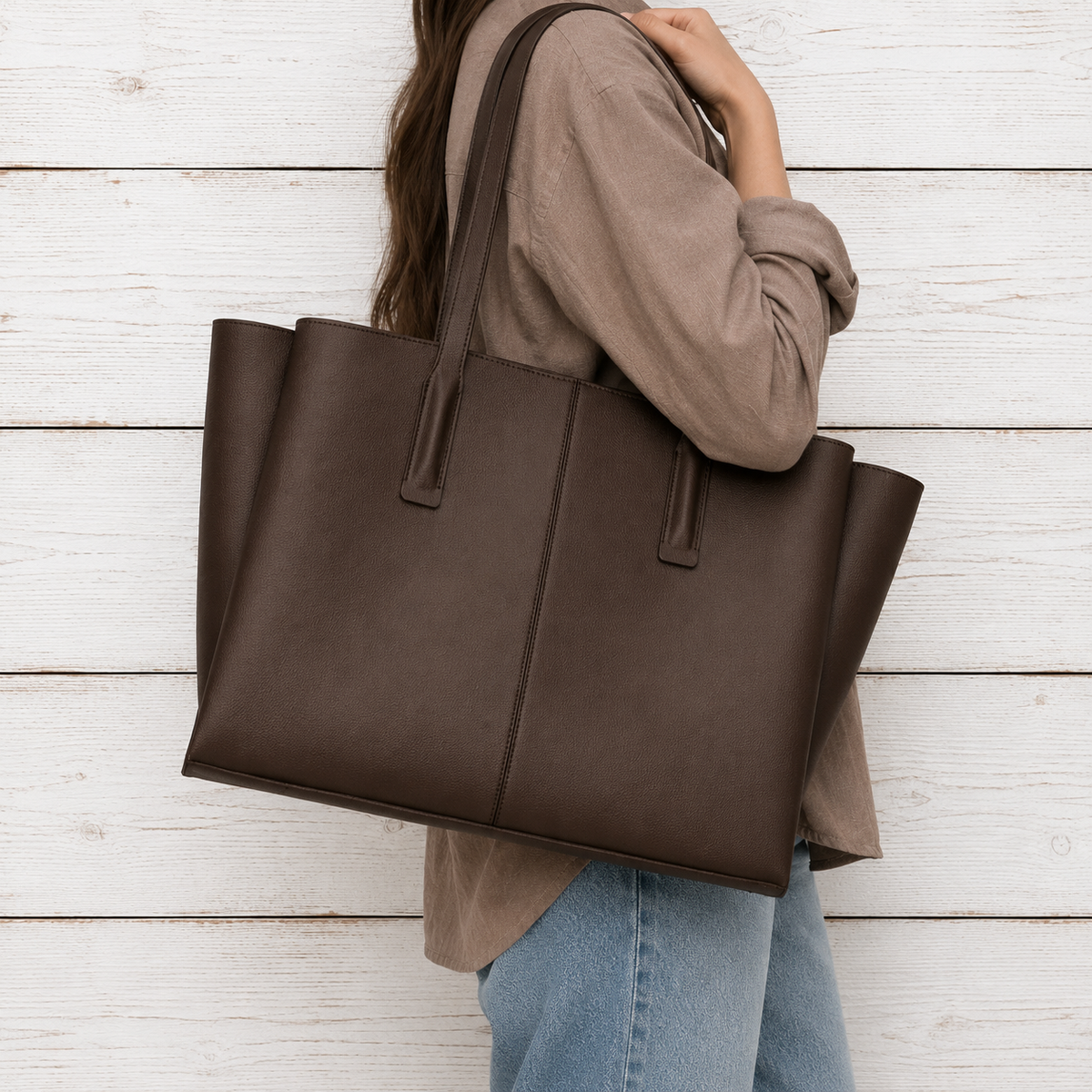 A person wearing a taupe long-sleeve shirt and blue jeans carries a large dark brown leather tote bag against a white wood plank wall.