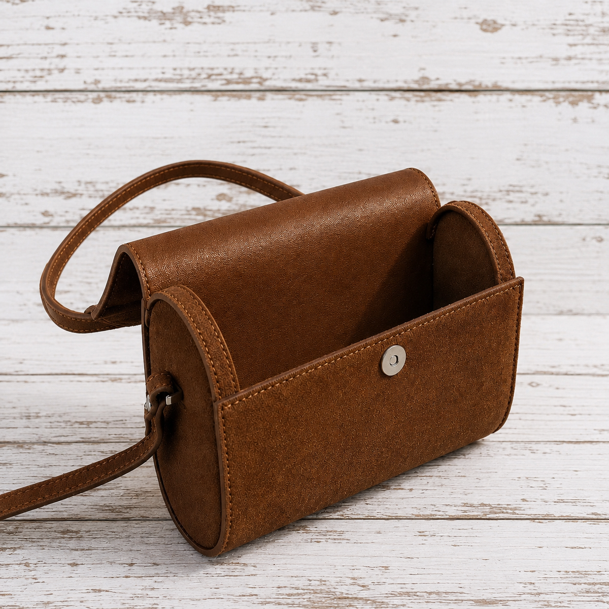 A brown faux-leather crossbody bag with a curved top handle and a silver snap button, displayed on a white distressed wood plank background.