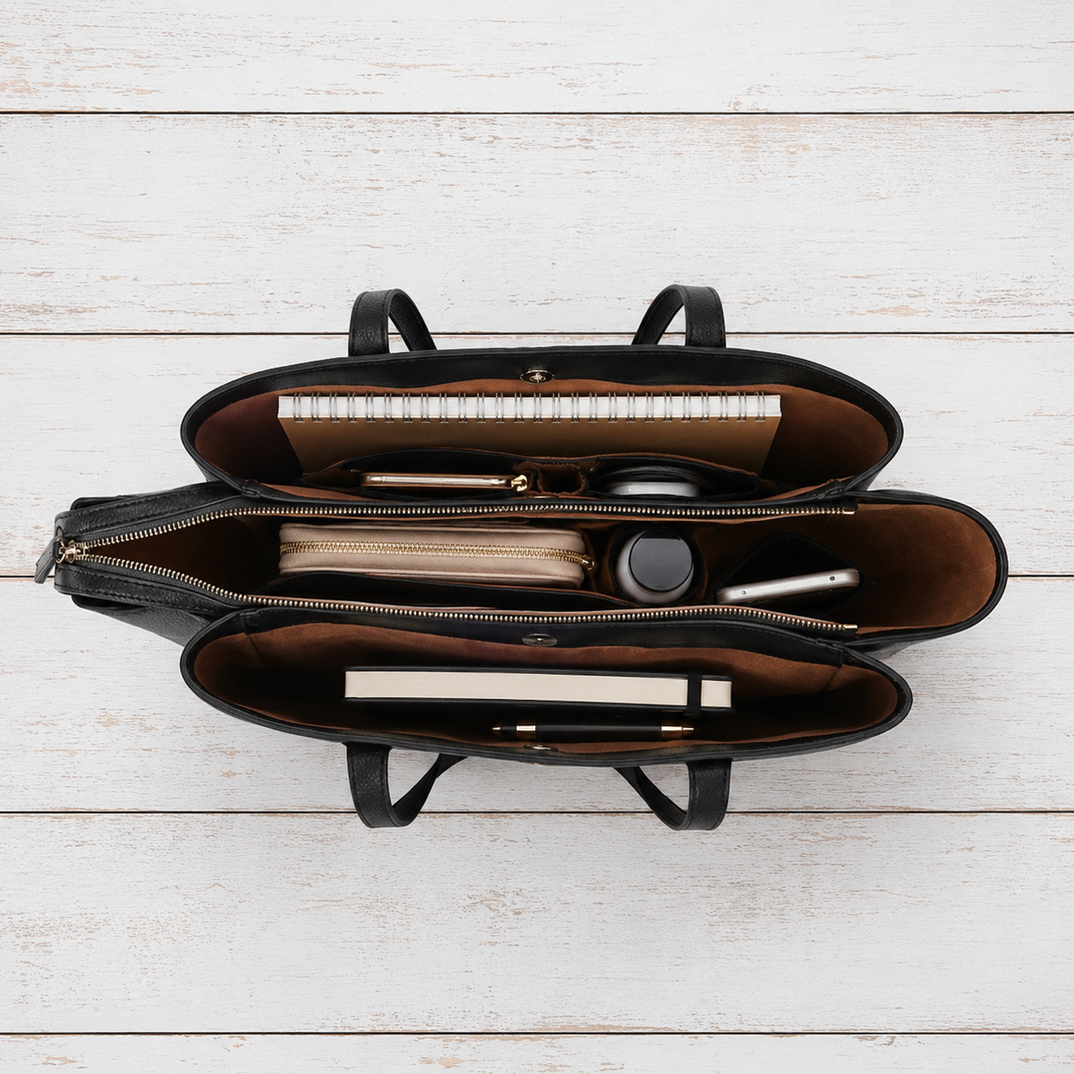 An open black leather organizer bag with brown interior lining, shown from a top-down view on a white wood plank background. Inside, a notebook, pens, a small pouch, and other accessories are neatly arranged.
