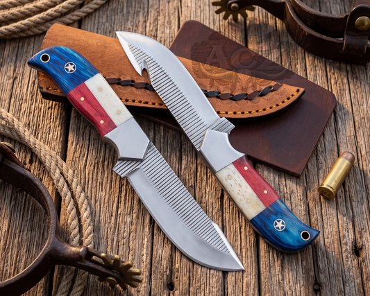 Two custom hunting knives with blue, red, and white handle scales are displayed on a rustic wooden surface alongside a brown leather sheath and western-themed props.