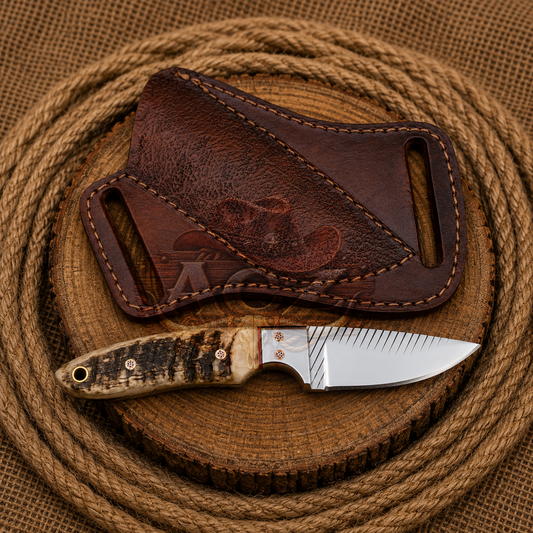 A fixed-blade knife with a carved bone handle and satin-finished steel blade rests on a round wooden slice, with a dark brown leather sheath behind it. The setup is photographed on a burlap surface with coiled rope in the background.
