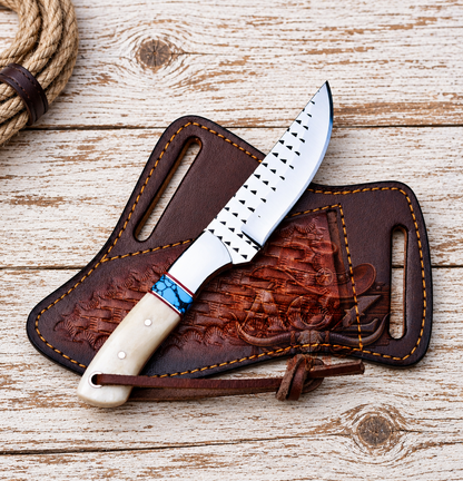A fixed-blade knife with a silver blade, black dotted guard, and cream handle with turquoise accents, resting on a brown embossed leather sheath over a rustic whitewashed wood background.