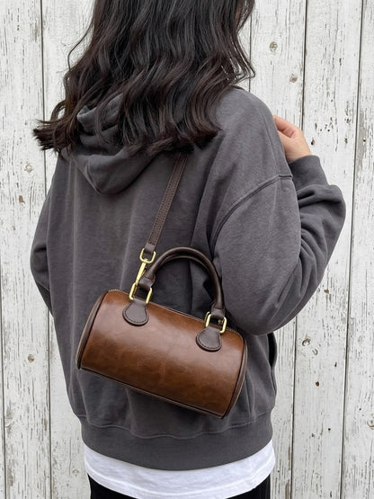 A person wearing a dark gray hoodie and white t-shirt is shown from behind, carrying a brown leather top-handle barrel bag with gold-tone hardware against a white wood plank wall.