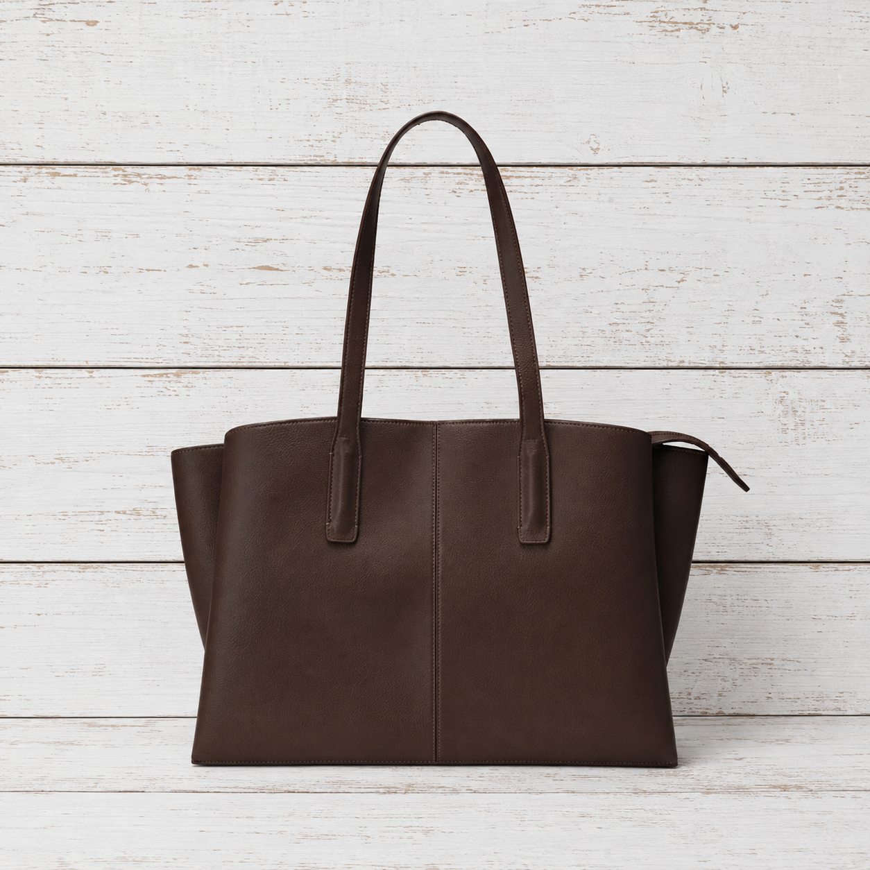 A dark brown leather tote bag with two shoulder straps, displayed against a white-washed wooden plank background.