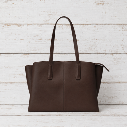 A dark brown leather tote bag with two shoulder straps, displayed against a white-washed wooden plank background.