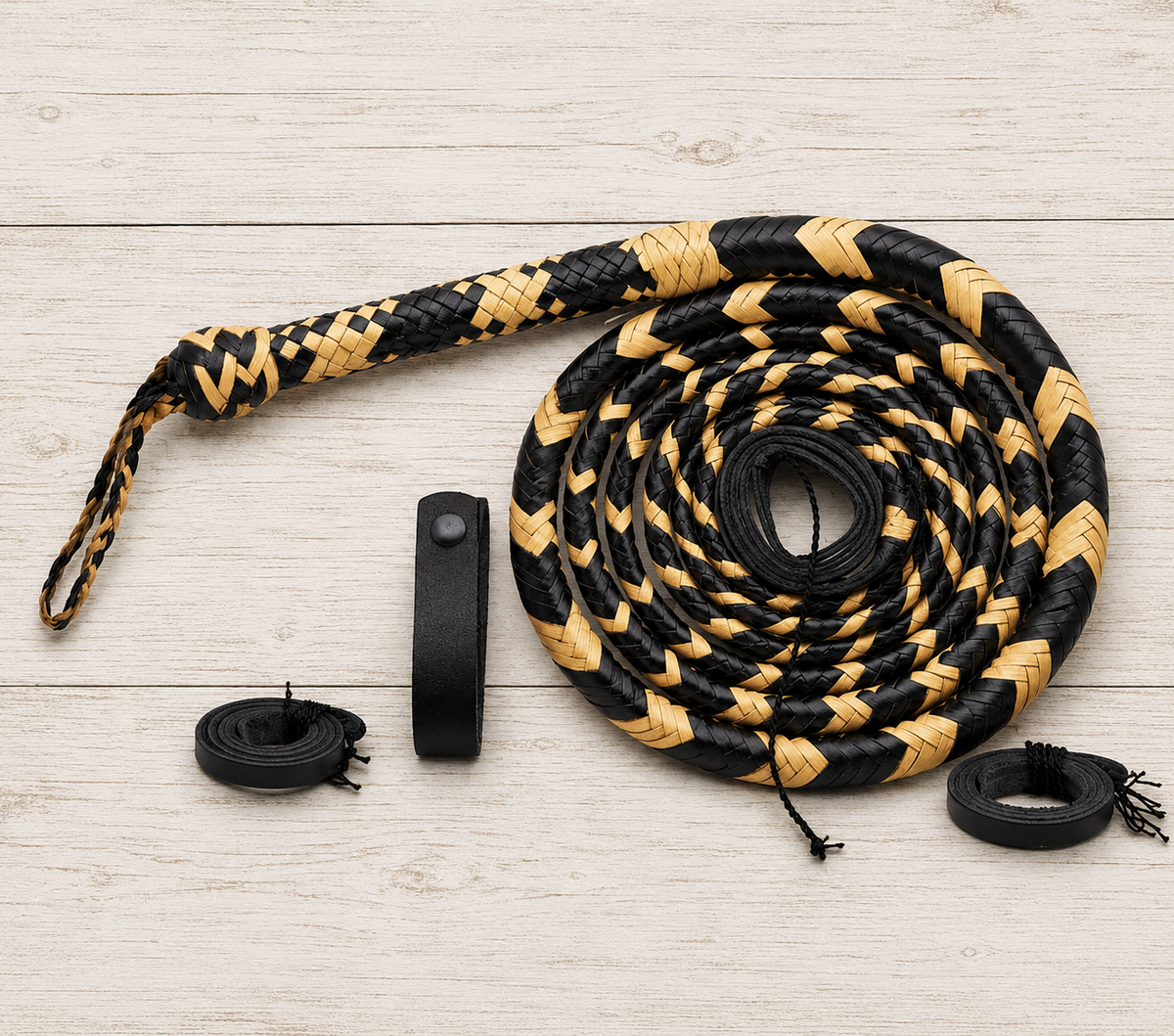 A coiled black and tan woven whip or flogger laid on a light wood plank background, with a black wrist strap and two small black rolled accessories placed beside it.
