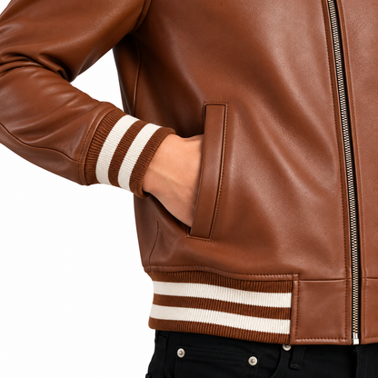 Close-up of a brown leather jacket with cream and brown striped ribbed cuffs and hem, shown on a model with a hand in the pocket against a white background.