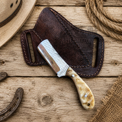 A fixed-blade knife with a rasp steel blade and a marbled bone handle rests on a dark brown leather sheath over a rustic wooden surface, with a hat, rope, horseshoe, and burlap visible around it.