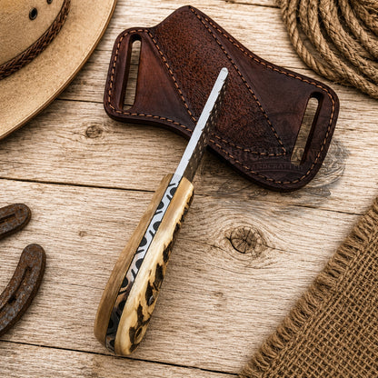 A handcrafted knife with a file steel handle and a dark steel blade rests diagonally on a rustic wooden surface next to its dark brown leather sheath. The scene is styled with western props including a straw hat, rope, and leather burlap.