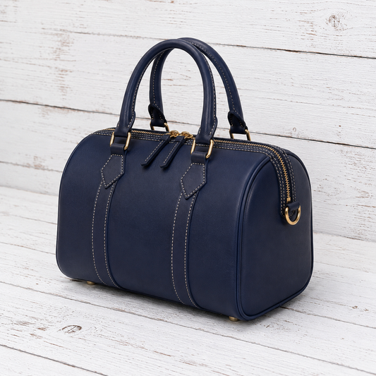 A navy blue leather handbag with gold-tone hardware, featuring dual top handles, decorative contrast stitching, and a zip-top closure