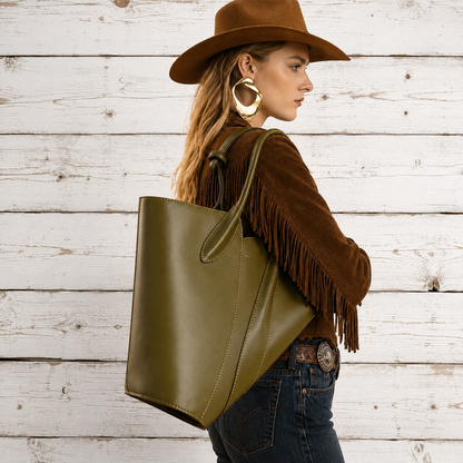 A woman in a brown hat and brown fringe jacket carries a large olive green leather tote bag 